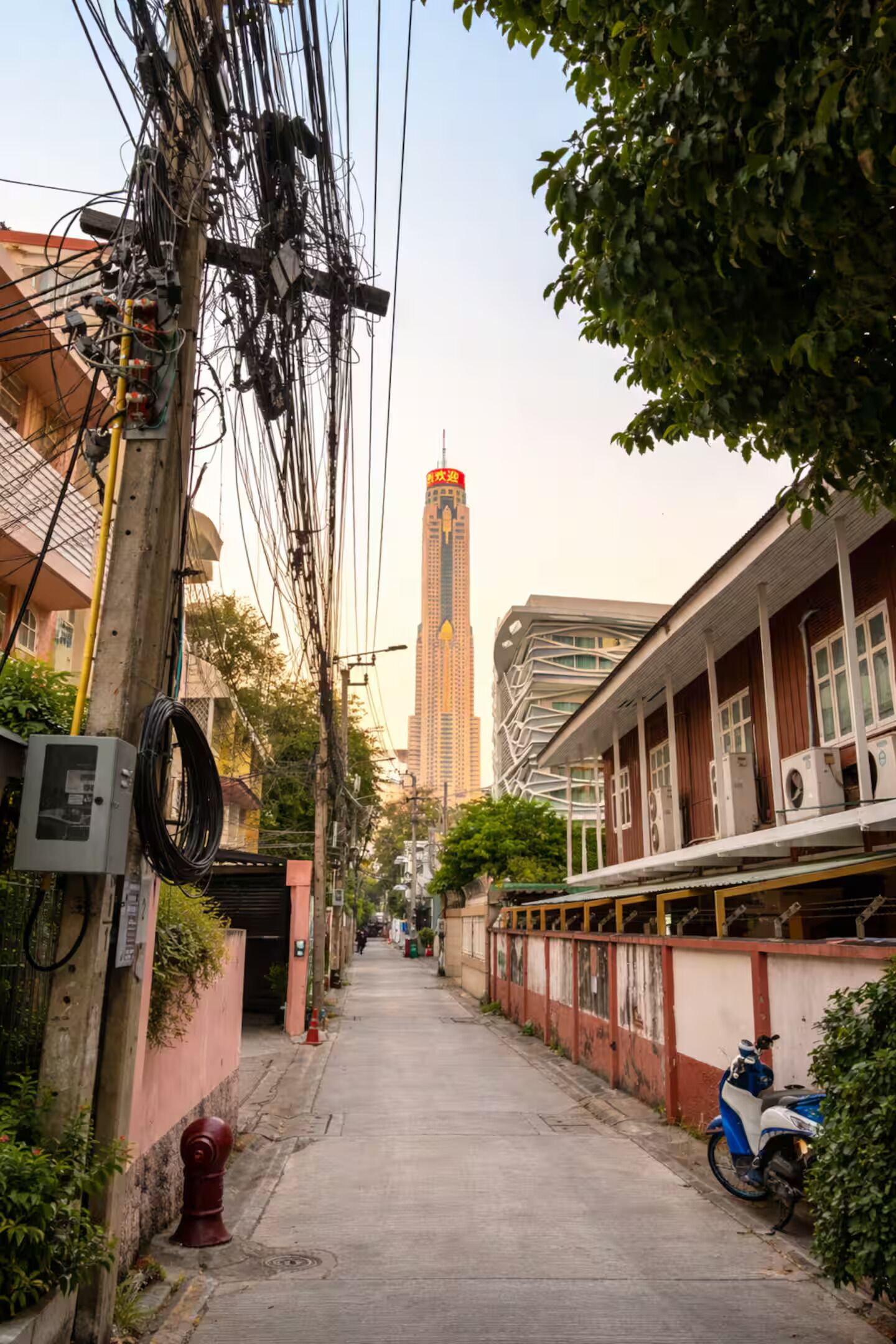 A narrow urban street with a building on the right, a tall skyscraper in the distance, and a parked scooter on the sidewalk.