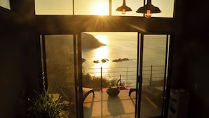 Una habitación con un gran ventanal que da a un balcón y vistas al mar al atardecer.