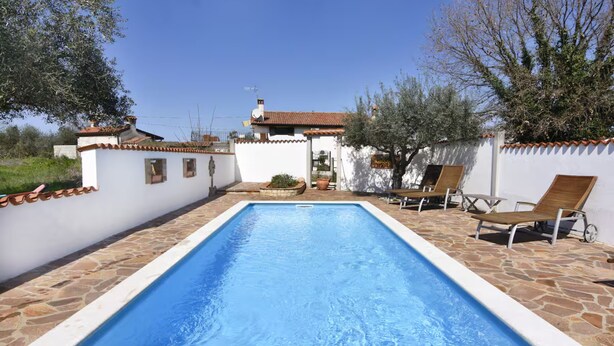 A rectangular swimming pool with a tiled deck, surrounded by white walls with terracotta tiles, and a small patio area with two wooden lounge chairs and a table. - Cozy Apartment With Pool Near Umag (Umag)
