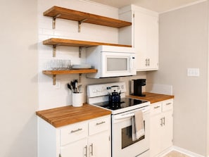 A kitchen with white cabinets, wooden shelves, a microwave, a stove, and a coffee maker.