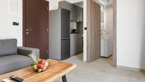 En moderne stue med en grå sofa, et træbord med frugt, et sort køleskab og et køkkenområde med grå skabe og en hvid bordplade.