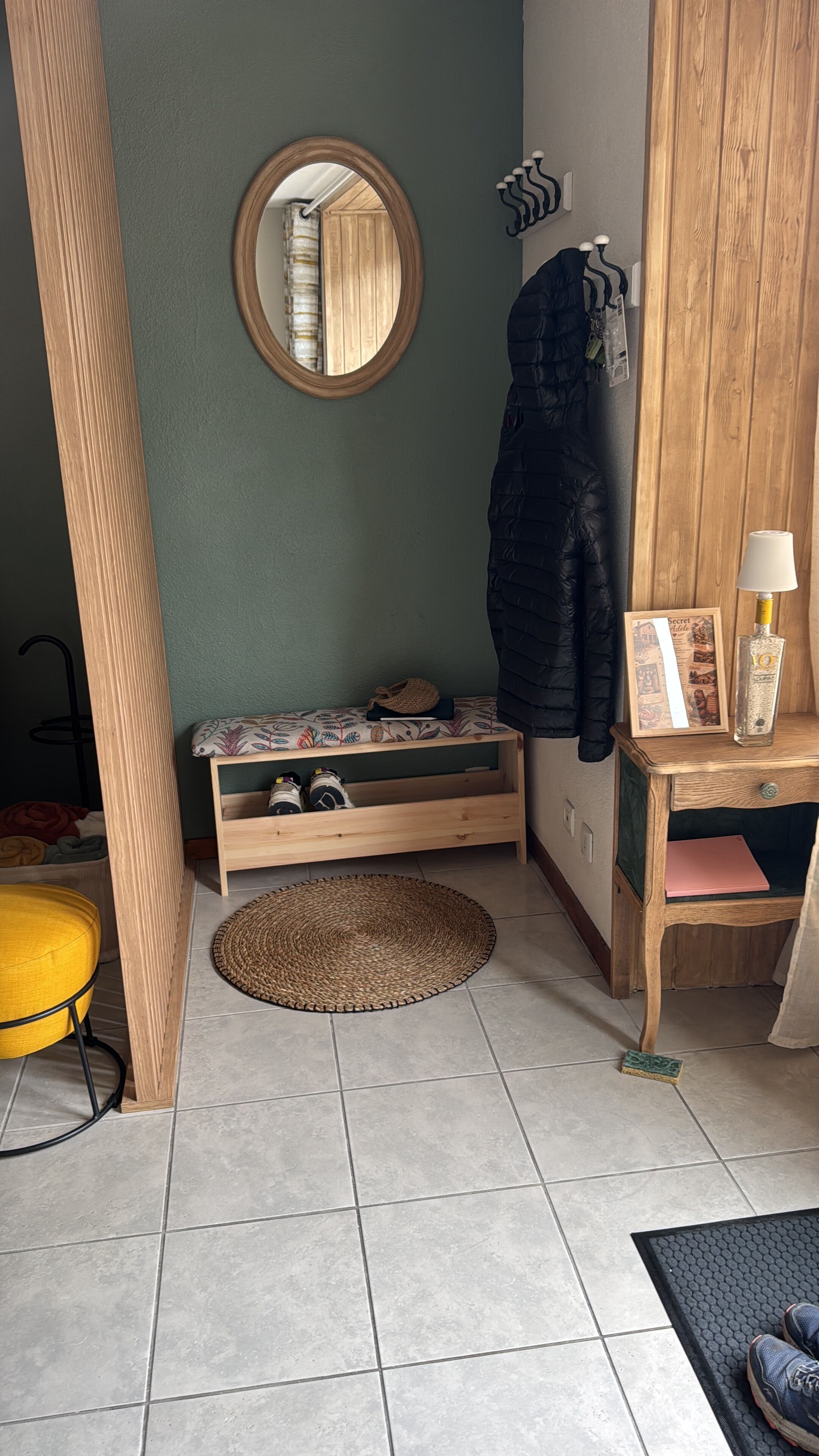 Une pièce comprenant un banc en bois, un miroir rond, un porte-manteau, une petite table avec une lampe et une porte dotée d’un cadre en bois.