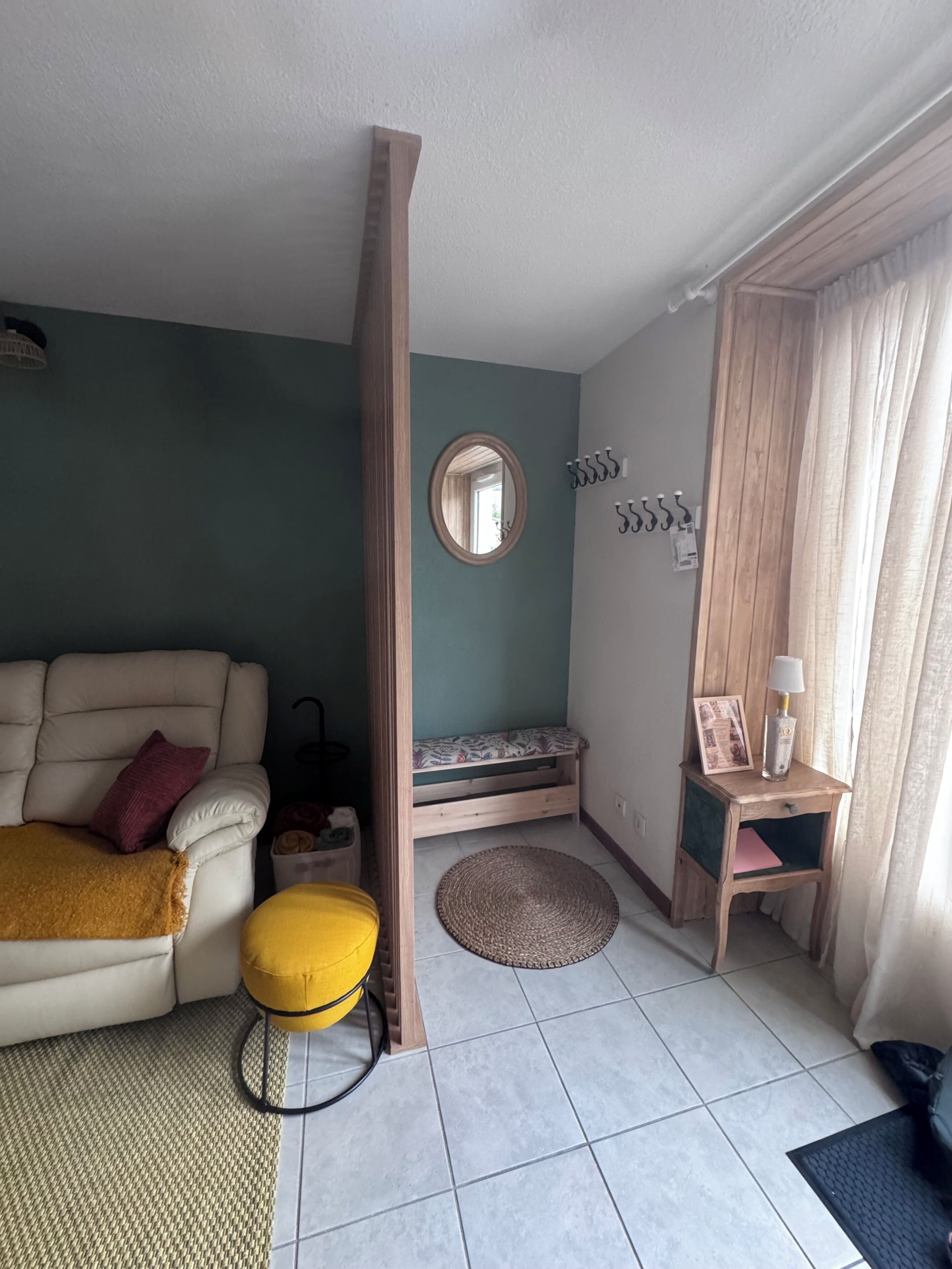Une pièce comprenant un canapé, un petit banc en bois, un miroir rond, un porte-manteau fixé au mur et une table de chevet avec une lampe et un cadre photo.