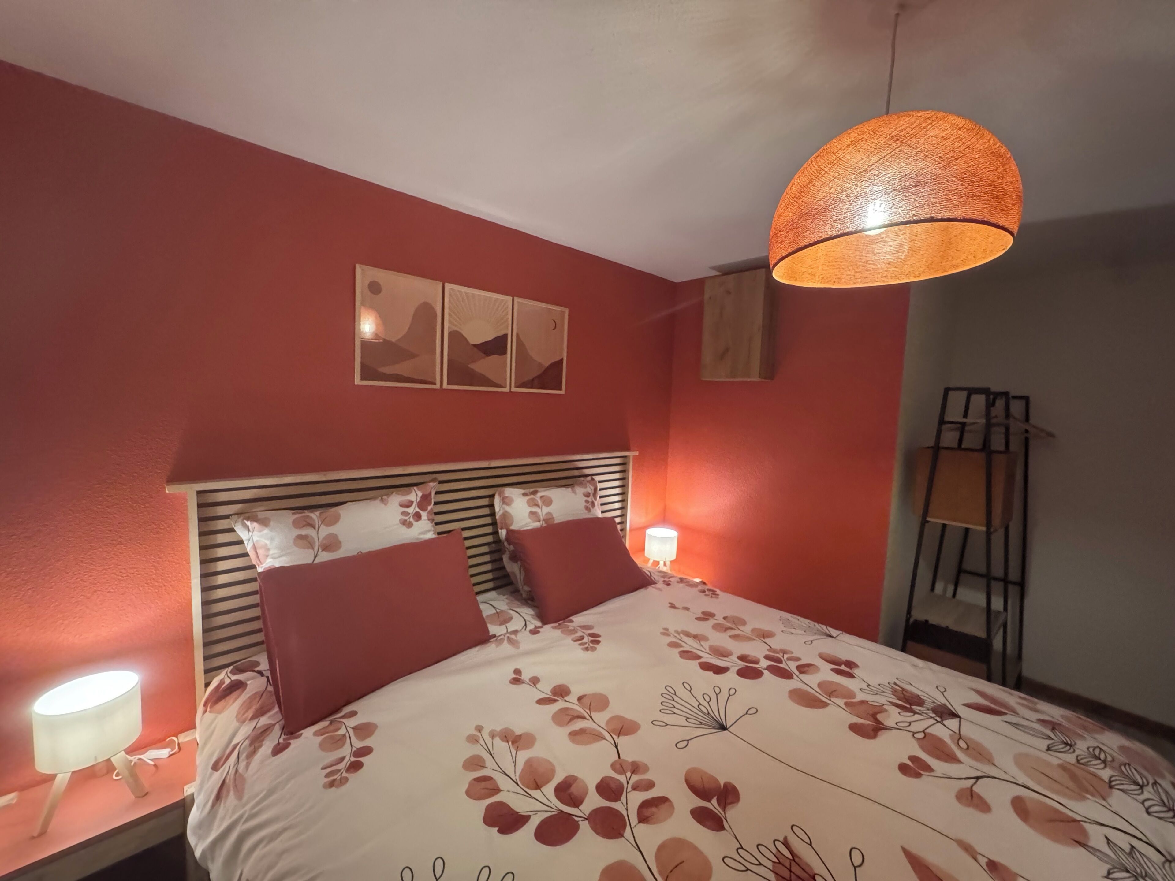 Une chambre à coucher avec un lit, deux lampes fixées au mur, un luminaire suspendu et une œuvre d’art encadrée au-dessus du lit.