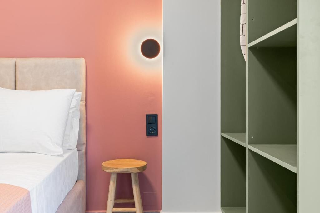 Un lit avec une literie blanche, un tabouret en bois, une lampe fixée au mur et une étagère verte.