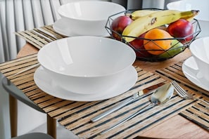 Une table à manger avec des assiettes blanches, un saladier rempli de fruits et un set de table en fibres tressées.