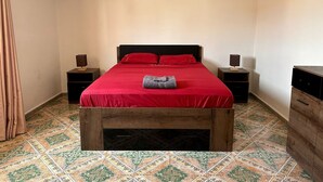 A bedroom with a wooden bed frame, red bedding, two bedside tables with lamps, and patterned floor tiles.
