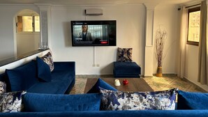 A living room with a large TV mounted on the wall, a blue sofa, a smaller blue armchair, and a coffee table with a vase and a red object.