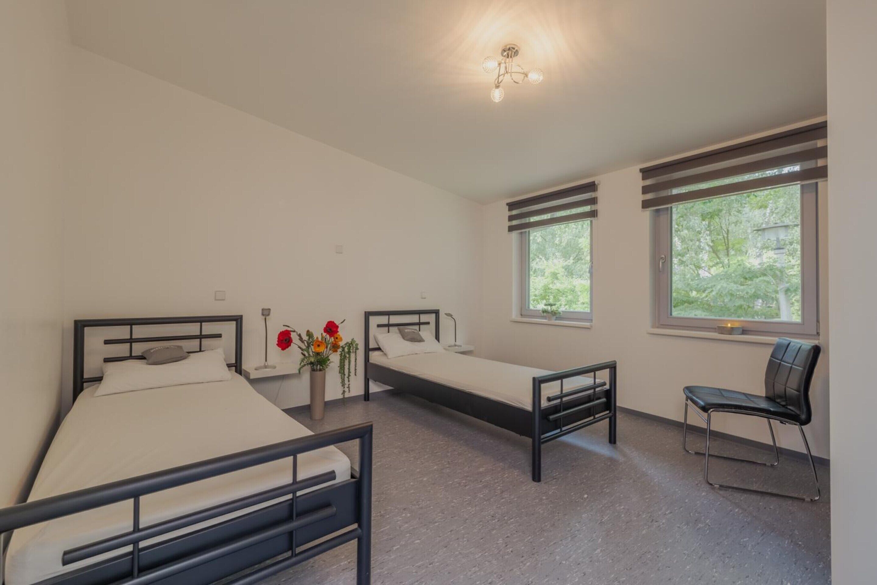 A room with two beds, a chair, a vase with flowers, and a window with a view of greenery.