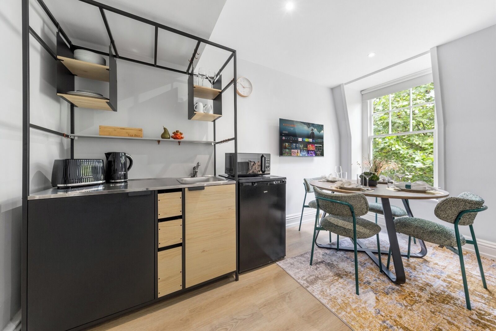 Une cuisine compacte aux couleurs noir et bois, un coin repas avec une table ronde et des chaises, et une grande fenêtre donnant sur un paysage verdoyant.
