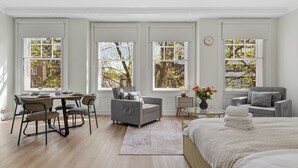 A spacious bedroom with a large bed, two grey sofas, a dining table with chairs, and three large windows with views of trees. - BAZ Tower - London (London)