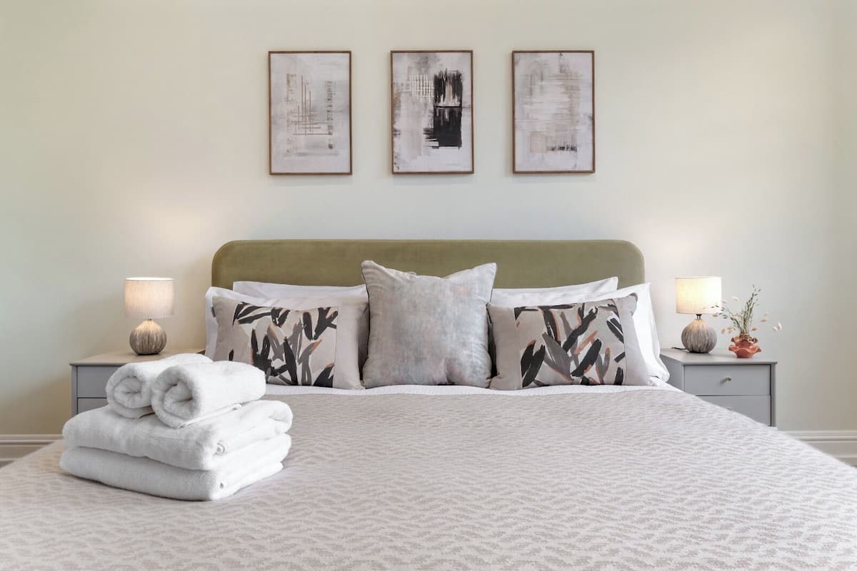 A neatly made bed with a patterned bedspread, a green upholstered headboard, and three framed artworks on the wall.