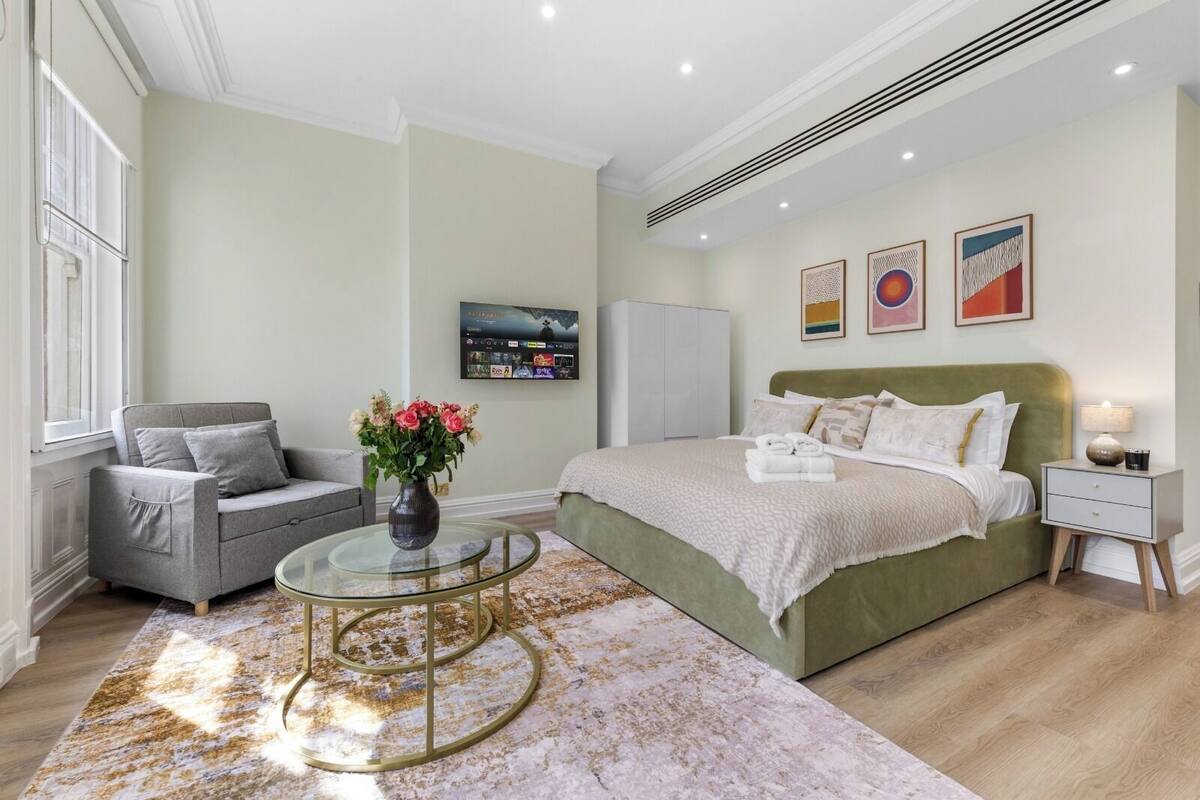 A modern bedroom with a green bed, a grey sofa, a glass coffee table, and a TV on the wall.