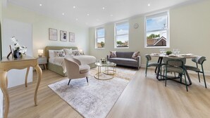 A modern hotel room with a bed, a sofa, a dining table with chairs, a small round table with a lamp, and large windows with a city view. - BAZ Tower - London (London)
