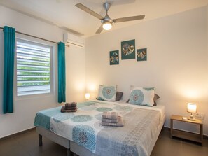 Ein Schlafzimmer mit Bett, Deckenventilator, einem Fenster mit Vorhängen und Wandbildern.