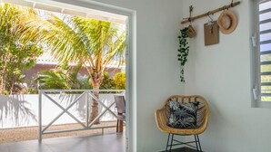 Ein Zimmer mit Blick auf Palmen, ein Korbstuhl mit gemustertem Sitzkissen, ein an der Wand befestigter Hutablage und eine weiße Tür.