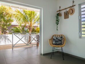 Ein Zimmer mit Blick auf Palmen, ein Korbstuhl mit gemustertem Sitzkissen, ein an der Wand befestigter Hutablage und eine weiße Tür.