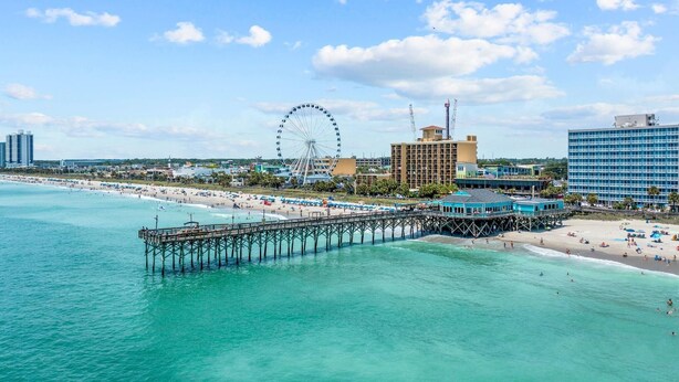 A coastal cityscape with a pier extending into turquoise waters, a large Ferris wheel, and a beachfront hotel. - 2-Bedroom Oceanfront Condo, Bay View Resort , 6th Floor, Indoor & Outdoor Pools (Myrtle Beach)