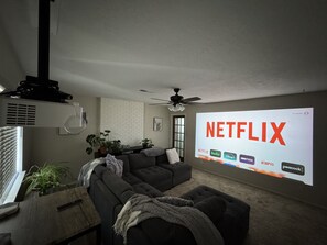 Ein Wohnzimmer mit einem großen Bildschirm, der das Netflix-Logo zeigt, einem Deckenventilator und einem modernen Design.
