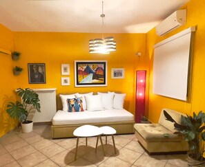 A room with a bed, a sofa, a small table, and a radiator. There are potted plants and artwork on the walls. - Alkmene Apartment - Warm Getaway in Kefalonia (Kefalonia)