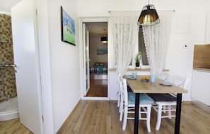 Un coin repas avec une table en bois, des chaises blanches, un luminaire suspendu et une vue sur la pièce adjacente.
