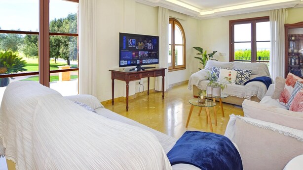 Een ruime woonkamer met een televisie, grote ramen, een bank en een bijzettafeltje.