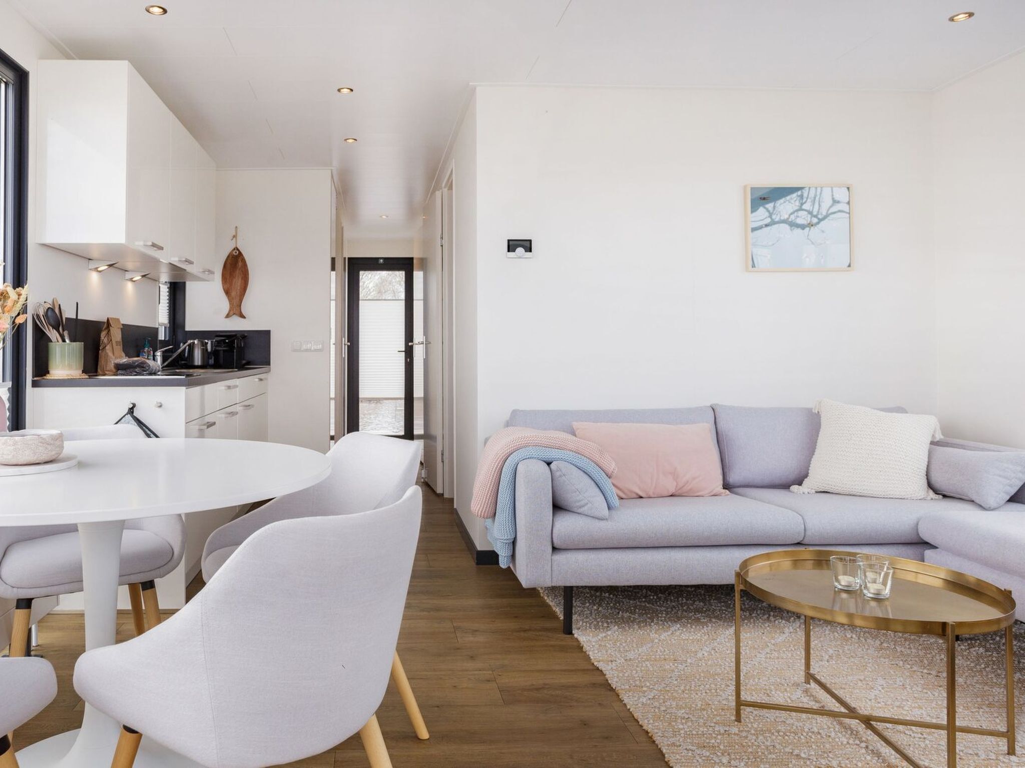 Un salon moderne avec un canapé gris, des chaises blanches, une table basse ronde et un coin cuisine doté de meubles blancs et d’un plan de travail noir.