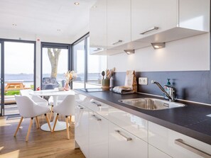A modern kitchen with a large window, a dining area with white chairs, and a view of the sea. - HouseBoat MeerRand by Interhome (Offingawier)