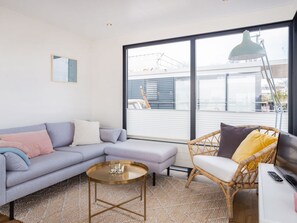 A modern living room with a sectional sofa, a round coffee table, a wicker chair, and a large window with a view of the city. - HouseBoat MeerRand by Interhome (Offingawier)