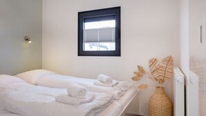 Et hotelværelse med to senge, et vindue med persienner, en lampe monteret på væggen og en dekorativ vævet kurv.