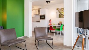 Ein modernes Wohnzimmer mit zwei Ledersesseln, einem Essbereich mit bunten Stühlen, einer Küche im Hintergrund und einem großen Flachbildfernseher an der Wand.