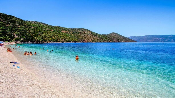 Una playa con aguas cristalinas, gente nadando y un paisaje montañoso al fondo.