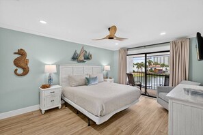 Ein Schlafzimmer mit Bett, Nachttischen, Deckenventilator, Wanddekoration mit Seepferdchen, Wandbild mit Segelboot, Balkon mit Blick und einem an der Wand befestigten Fernseher.