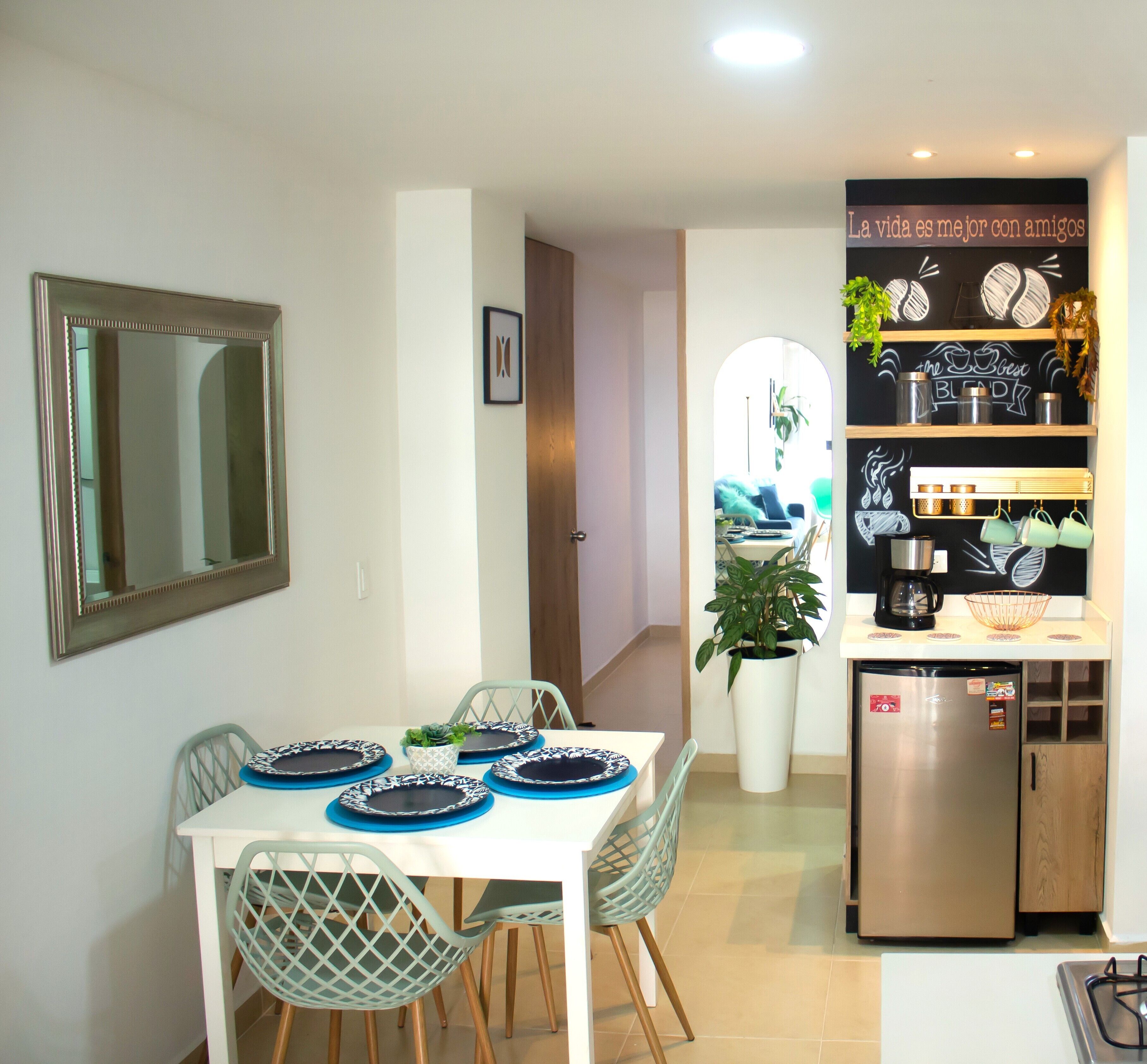A dining area with a white table, blue plates, and green chairs. A kitchen with a refrigerator and shelves is visible in the background.