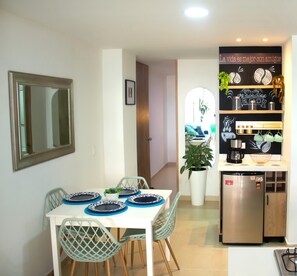 A dining area with a white table, blue plates, and green chairs. A kitchen with a refrigerator and shelves is visible in the background. - Adorable 1-bedroom apartment in a charming setting, perfect for relaxing (Sabaneta)