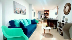 A modern living room with a blue sofa, teal armchairs, a coffee table, and a kitchen area in the background.