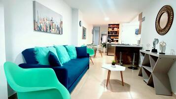 A modern living room with a blue sofa, teal armchairs, a coffee table, and a kitchen area in the background.