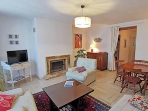 Ein Wohnzimmer mit Kamin, einem Fernseher auf einem weißen Schrank, einem Esstisch mit Stühlen und einer hängenden Leuchte.
