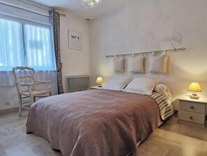 Ein Schlafzimmer mit einem Bett, zwei Nachttischen mit Lampen, einem Sessel, einem Heizkörper und einem Fenster mit Vorhängen.