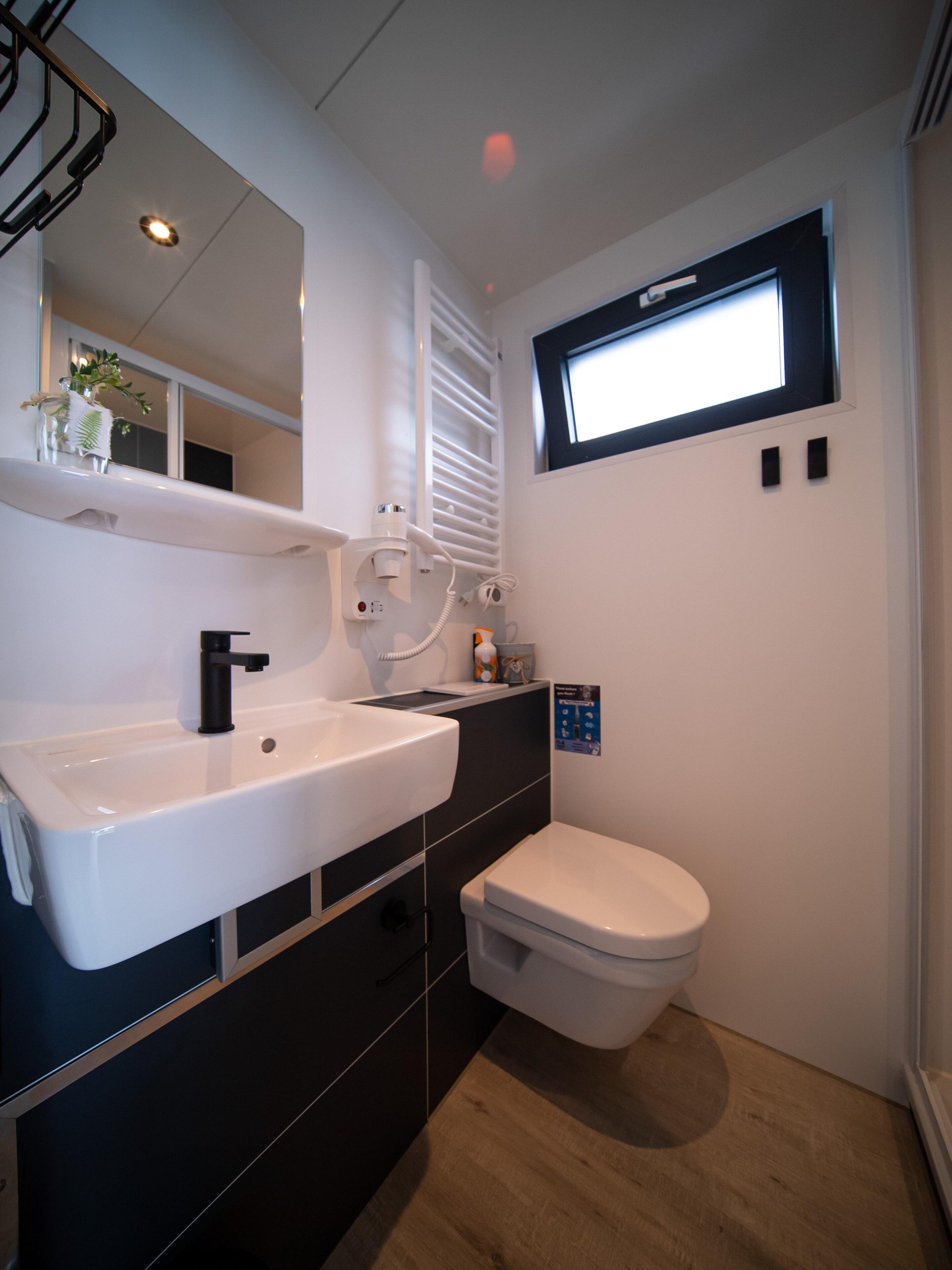 A compact bathroom with a white sink, black cabinets, a white toilet, and a window with blinds.