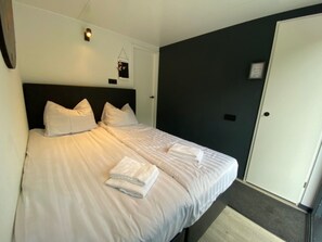 Una cama bien hecha con almohadas y toallas dobladas, una pared oscura y una puerta.