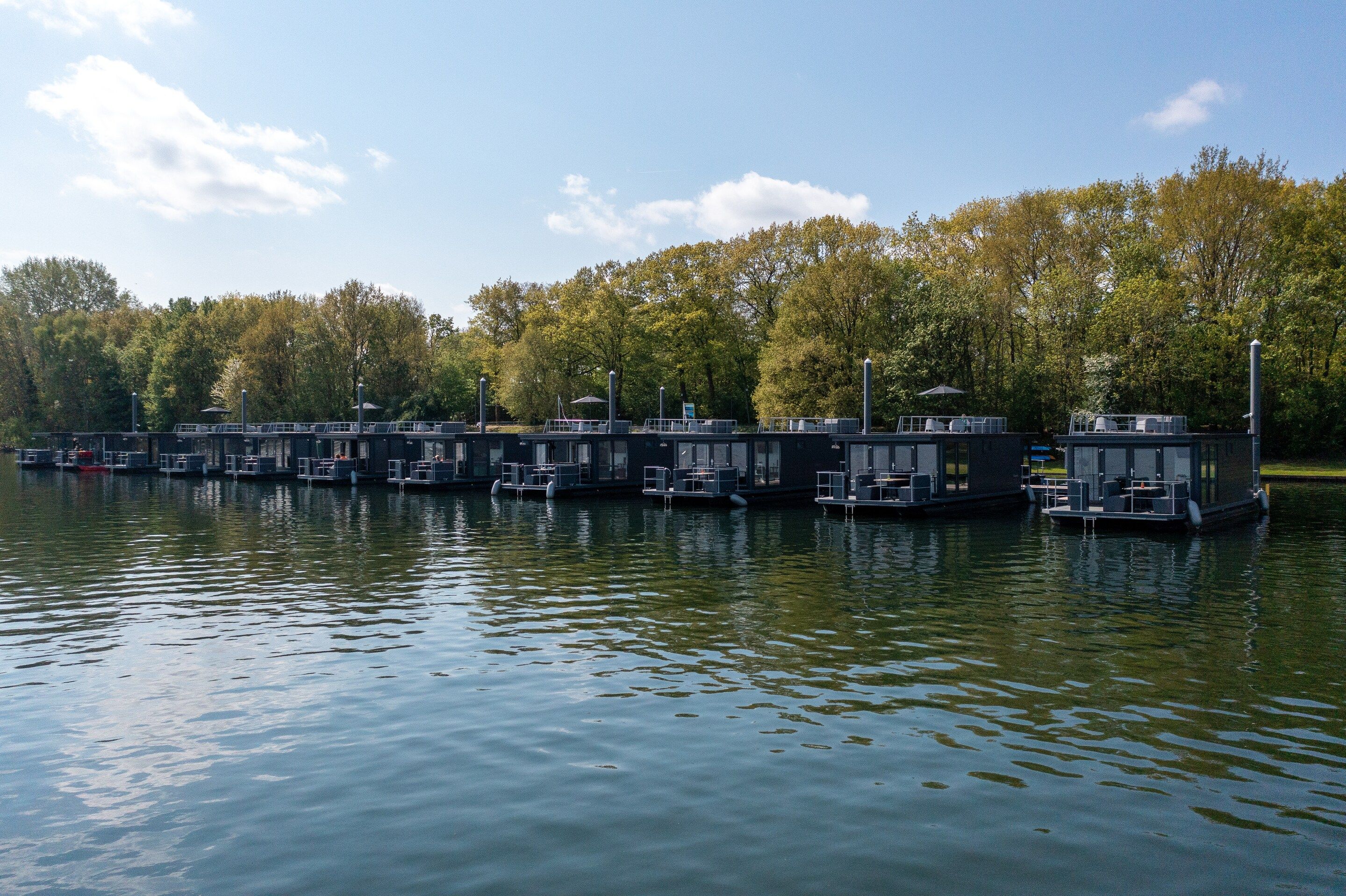 A row of floating structures with outdoor seating areas, connected by walkways, set against a backdrop of trees and a clear sky.