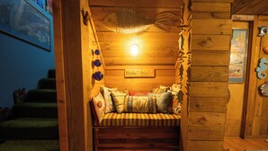 Une pièce chaleureuse, aux murs en bois, avec un banc, une enseigne indiquant « Antre de Hobbit », et un motif de poisson décorant le plafond.