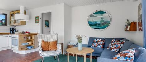 A modern living room with a sofa, armchair, and coffee table. There is a kitchen area with cabinets and a stove.