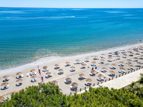 A beach with rows of sun loungers and umbrellas, a clear view of the sea, and a rocky shoreline.. Marinez Hotel – All Inclusive