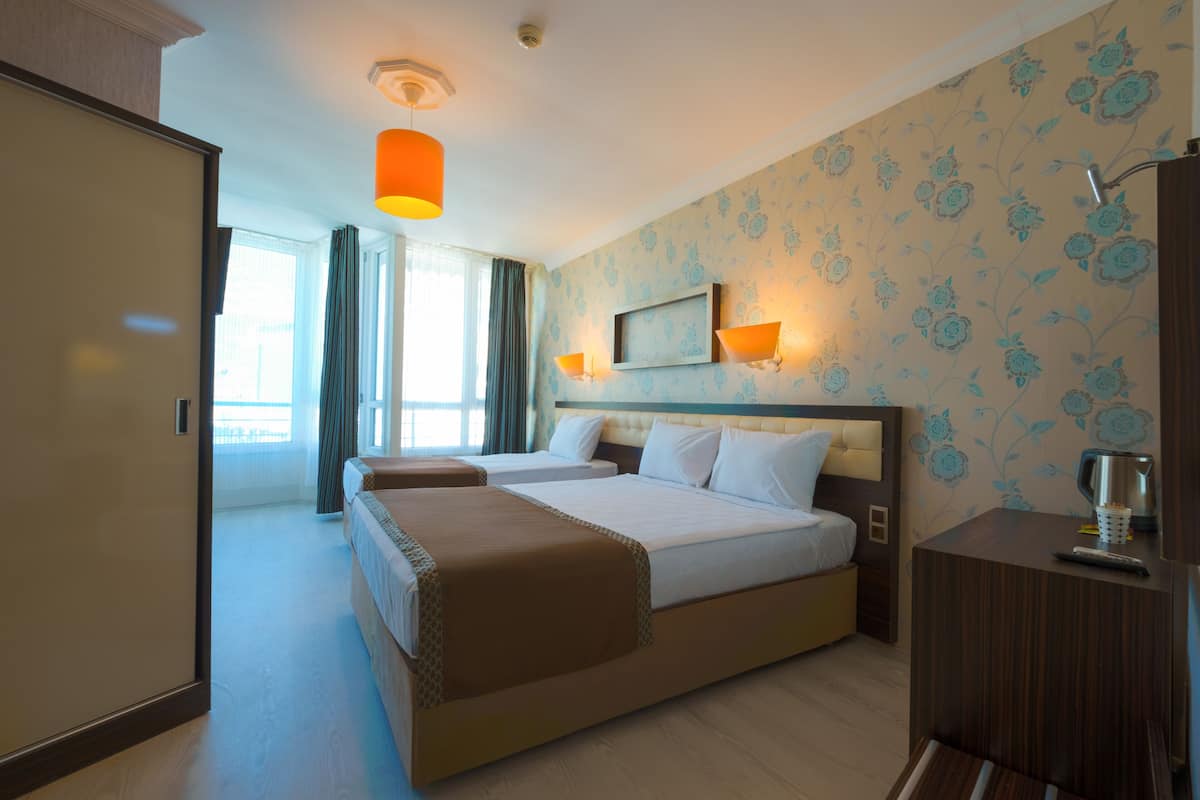 Ein Hotelzimmer mit zwei Betten, einem Holz-Kopfstück, einem Nachttisch mit Tasse und Fernbedienung sowie einem Fenster mit Vorhängen.