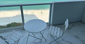 Un balcón con vista a la playa, una mesa redonda y dos sillones blancos.