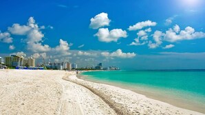 Ein Strand mit blauem Himmel, sandigem Ufer und Gebäuden im Hintergrund.