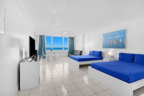 A hotel room with a bed, a desk, a chair, a table, and a view of the sea. - 530 - Large Studio Ocean View (Miami Beach)