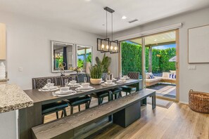 Un espace de repas moderne avec une longue table, plusieurs chaises et un banc. Une grande fenêtre offre une vue sur la verdure à l’extérieur.